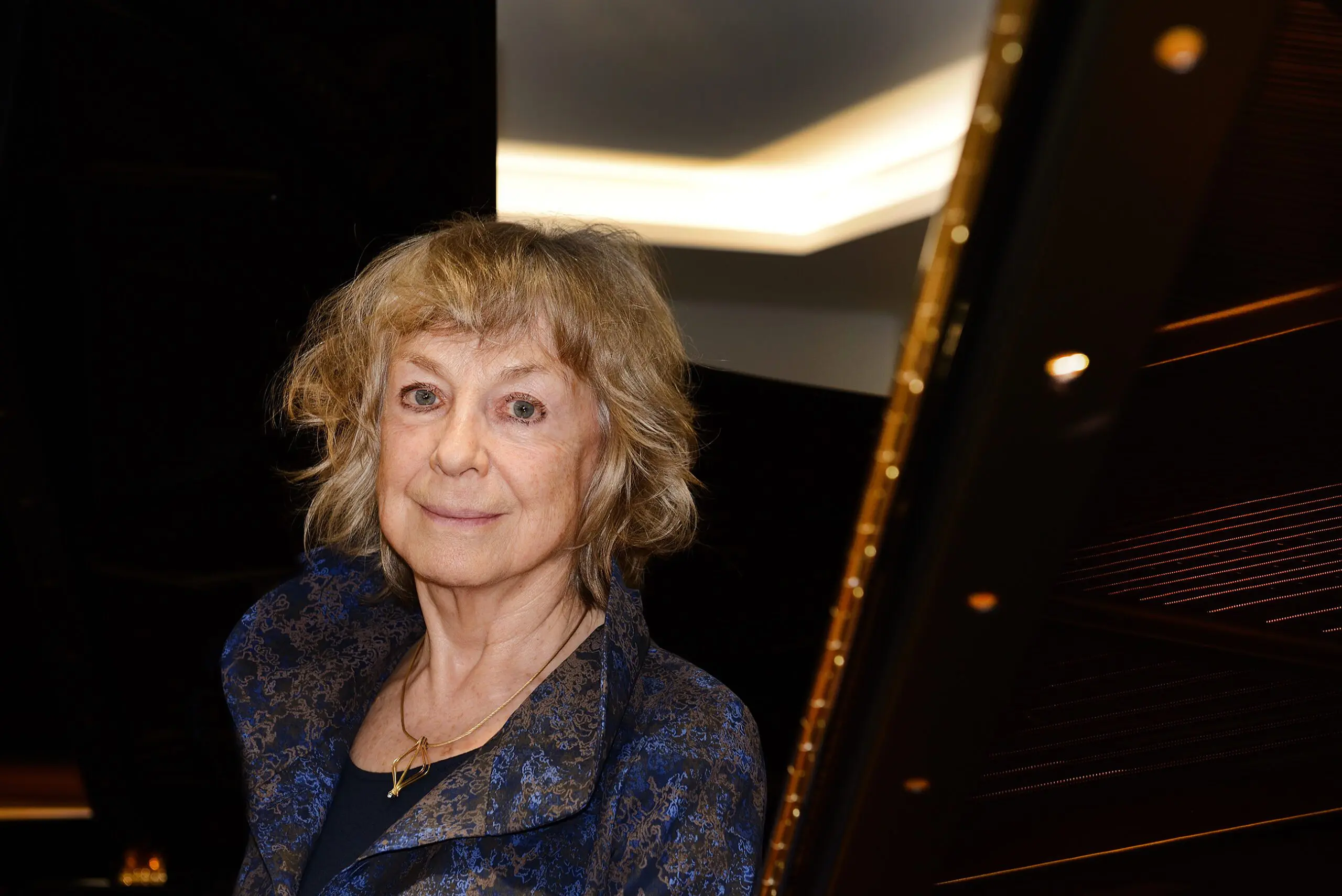 Portrait von Marianne Schroeder. Ältere blonde Frau mit dunkelblauem Oberteil vor einem Flügel